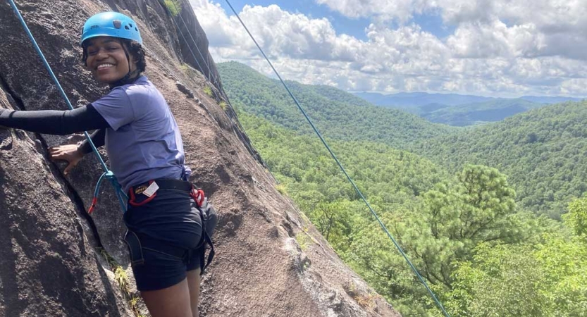 A person wearing safety gear is secured by ropes as they pause climbing a rock wall to smile for the photo.